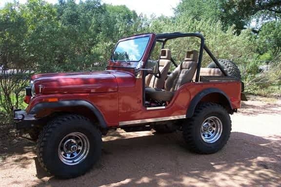 A red 1978 Jeep CJ5 photographed from the side with the top off, showing the tan interior, roll bar, spare tire and chrome wheels, parked on dirt among trees
