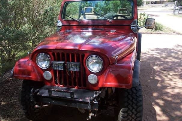 A red 1978 Jeep CJ5 photographed from the front three-quarter on a dirt drive, with the top off, chrome wheels, knobbly tires and auxiliary lights in the grille
