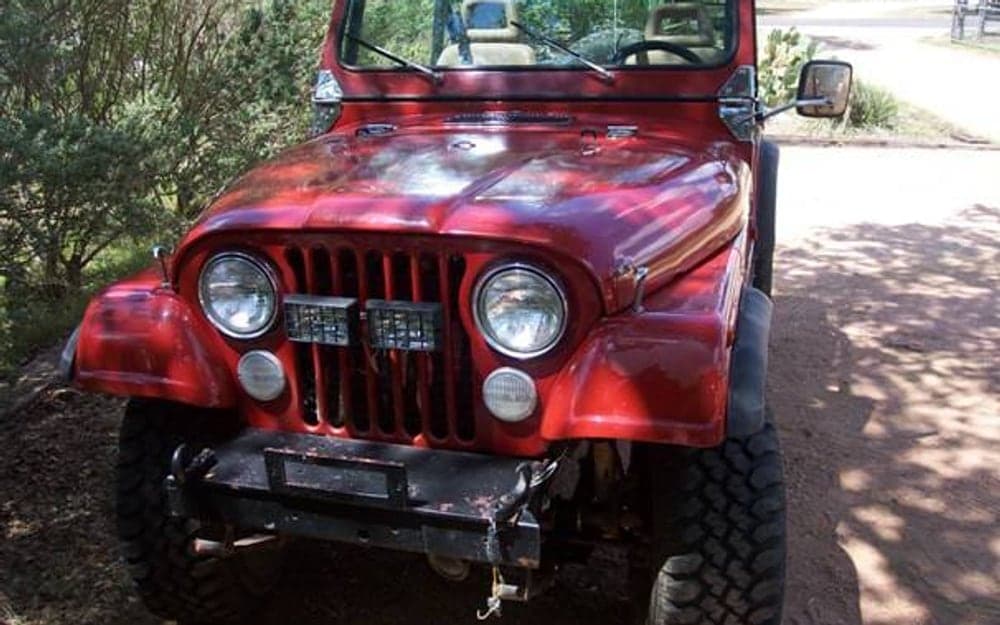 A red 1978 Jeep CJ5 with the top off, photographed from the front three-quarter on a dirt drive, on chrome wheels with knobbly tires