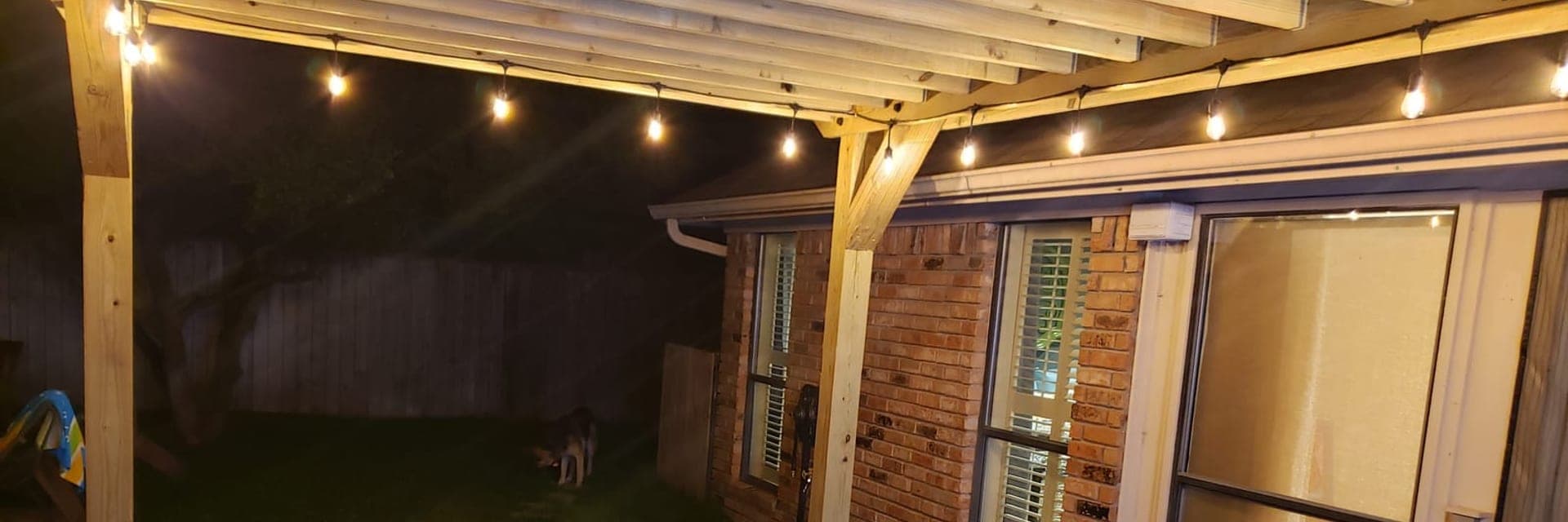 A cedar pergola over a patio at night, strung with warm bulb lights, seen from underneath