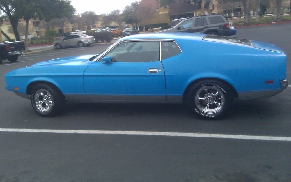 A blue 1971 Ford Mustang Mach 1 fastback photographed from the side in a parking lot, on chrome five-spoke wheels with raised-white-letter tires