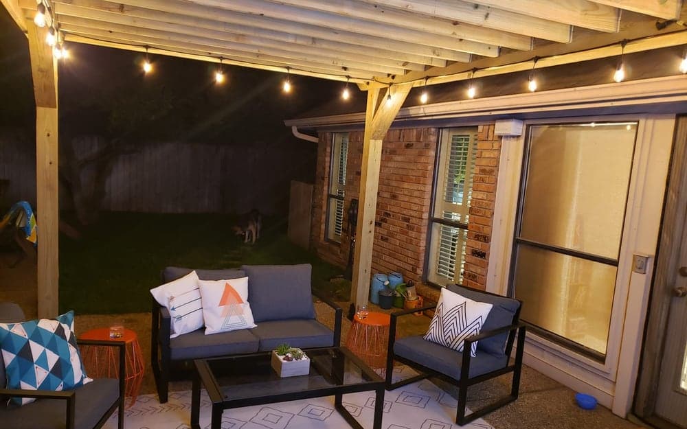 A cedar pergola over a patio at night, strung with warm bulb lights, with outdoor furniture underneath and a brick house wall behind