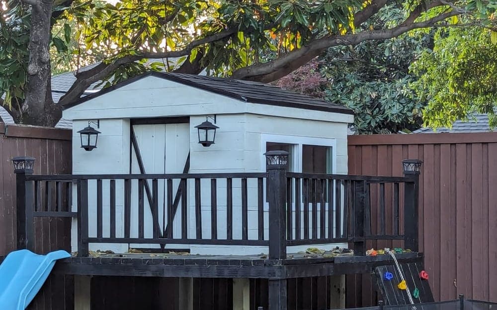 A white-painted elevated playhouse with a black shingled roof and a dark stained deck and railing, standing on wooden posts in a fenced backyard corner