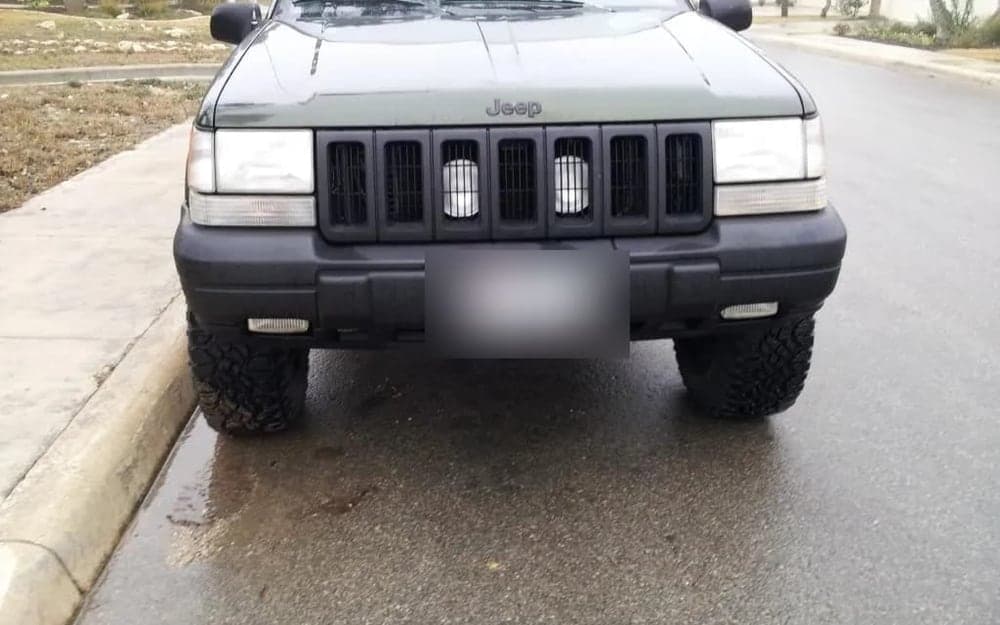 A dark green 1996 Jeep Grand Cherokee ZJ photographed head-on, lifted on black steel wheels and all-terrain tires, with auxiliary lights fitted behind the grille slots